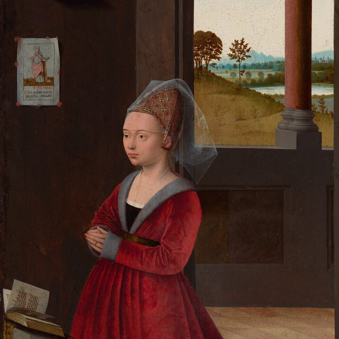 A pale-skinned woman wearing a voluminous, garnet-red, long-sleeved dress kneels at a low lectern in a dark gray room, in front of a window opening onto a grassy landscape in this vertical painting. The woman’s body is angled to our left, and she looks off in that direction. She has a high forehead, thin, arched brows, light brown eyes, a round jawline, and her pale lips are closed. A cone-shaped, gold headdress covers her hair. On the headdress, pearls mark the intersections of a grid made by lines of white dots, and there is a red flower in the center of each square. A gauzy, translucent veil covers her forehead to her brows and hangs off the back of the headdress. Another sheer cloth covers the woman’s chest down to the neckline of her dress, which is edged with gray fur. A gold-colored belt gathers the full skirt, which pools on the ground around and behind her. Gray cuffs cover the backs of her hands to her fingers, which touch in prayer in front of her waist. She wears a gold ring on the ring finger we can see. The gray lectern at which she kneels is cut off by the left side of the painting. Pages flutter across an open book laid on the angled top of the lectern.